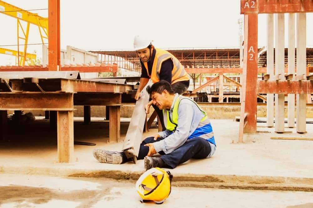 Injured construction worker holding his leg while a coworker helps at a job site