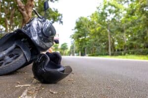 A black motorcycle on its side near a black helmet lying on the asphalt road, surrounded by greenery and trees